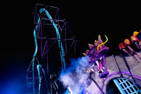  Atracción de montaña rusa iluminada de noche en un parque de diversiones. En primer plano se ve la escultura de un dragón morado que expulsa humo, mientras detrás se eleva la estructura metálica de la montaña rusa con rieles iluminados contra el cielo oscuro.