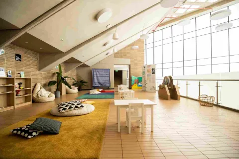  Espacio interior luminoso acondicionado para actividades infantiles, con mesas y sillas pequeñas, cojines sobre alfombras y estanterías con libros y materiales. El lugar cuenta con grandes ventanales que dejan entrar luz natural y paredes de ladrillo que crean un ambiente cálido y educativo.