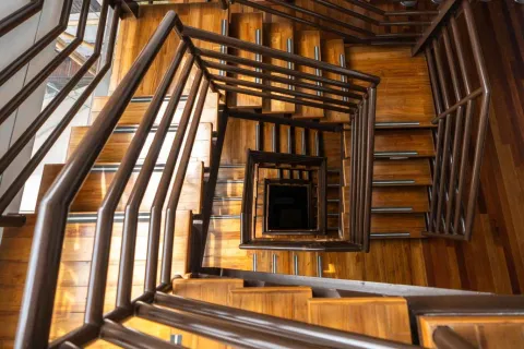 Vista hacia abajo de una escalera interior de madera con barandas metálicas oscuras que forman una figura geométrica en espiral hacia el centro. Los peldaños de madera iluminados por luz natural crean un patrón cálido y simétrico que desciende varios niveles del edificio.
