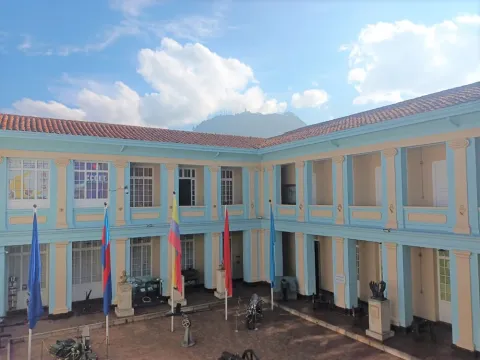  Patio interior de un edificio histórico de dos pisos con columnas y balcones, banderas izadas en el centro y piezas militares exhibidas, con montañas visibles al fondo.