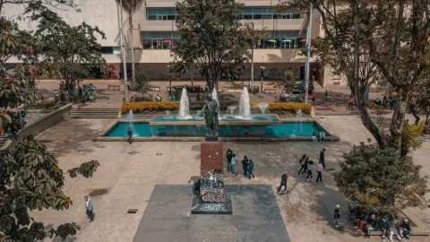 Vista elevada de una plaza urbana con una estatua central sobre pedestal, rodeada por una fuente rectangular con chorros de agua. Varias personas caminan y se reúnen en el espacio, mientras árboles y zonas verdes delimitan el lugar. Al fondo se observa un edificio de varios niveles, y el ambiente refleja actividad cotidiana en un espacio público durante el día.