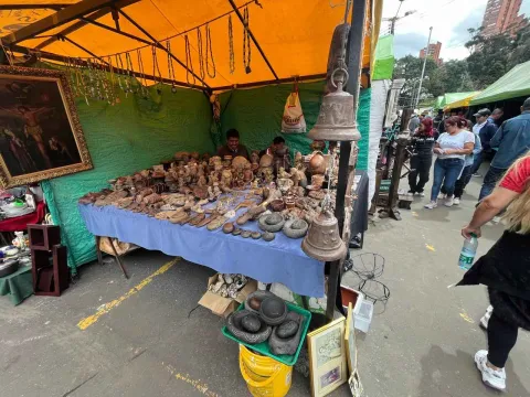 Puesto de mercado al aire libre bajo una carpa amarilla, donde se exhiben objetos antiguos y artesanales como figuras, utensilios, piedras y piezas decorativas sobre una mesa cubierta. Al fondo hay vendedores atendiendo y, a un lado, visitantes recorren el lugar.