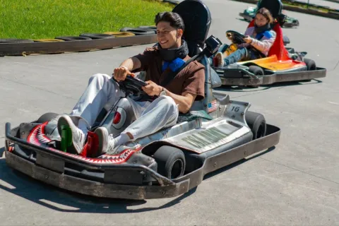 Dos personas conducen karts en una pista al aire libre en un parque de diversiones. En primer plano, un joven sonríe mientras maneja su kart gris, seguido por otra persona en un kart naranja. Alrededor de la pista se observan llantas de protección y césped verde.