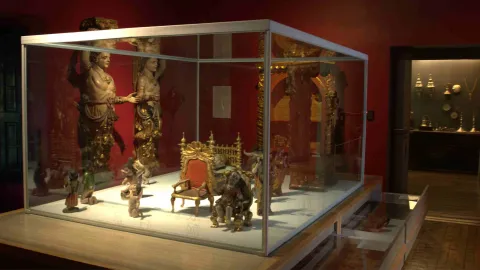 Museum display case with antique sculptures and decorative objects, including small figures, a miniature throne, and carved pieces, in a dimly lit room with red walls.