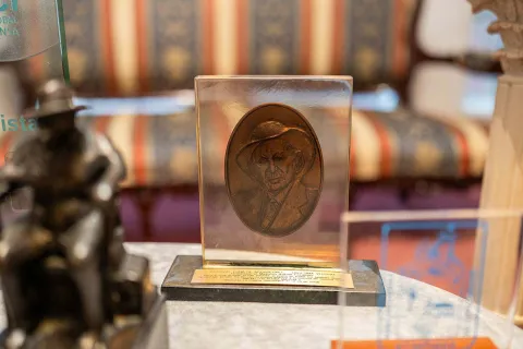 Commemorative object displayed in a glass case, consisting of a plaque with a relief of a male face on a base with an inscription. The piece has a gold finish and is surrounded by other decorative elements and awards, in an interior setting reminiscent of a museum or exhibition hall.