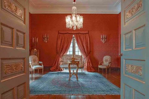  Vista de un elegante salón de estilo clásico en tonos rojos, con paredes decoradas y cortinas pesadas enmarcando una ventana central. En el centro hay una mesa de madera tallada sobre una alfombra azul, rodeada de sillas tapizadas de estilo antiguo. Un candelabro de cristal cuelga del techo, y la escena se observa desde una puerta abierta que enmarca el espacio con detalles ornamentales.