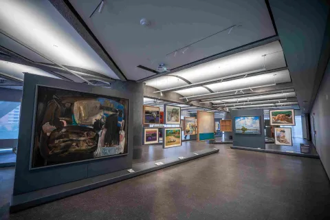 Interior view of the Bancolombia Art Gallery, a modern exhibition space showcasing various large-format paintings and framed works on panels and walls. The spacious room, with lighting directed from the ceiling, allows visitors to explore the exhibition and appreciate the works from different angles in a contemporary setting.