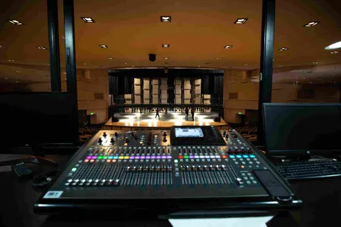  Vista desde la cabina técnica de un teatro, donde se observa una consola de sonido con botones y luces de colores en primer plano. Al fondo se ve el escenario iluminado y parte del auditorio con filas de asientos, mientras algunas personas caminan sobre el escenario durante un ensayo o preparación técnica.
