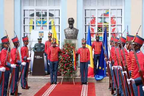 Ceremonia militar con guardia de honor uniformada frente a un busto conmemorativo, acompañada por banderas y una ofrenda floral en el patio de un edificio histórico.