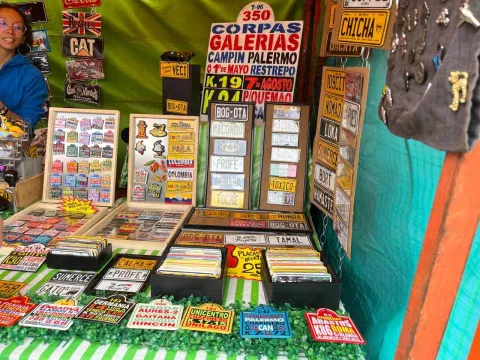Puesto de mercado con exhibición de placas decorativas y souvenirs coloridos, inspirados en nombres, lugares y expresiones populares. Los objetos están organizados en mesas y paneles verticales, con letreros llamativos de oferta.