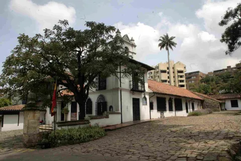 White colonial house with red tile roofs and a small bell tower, surrounded by trees and a cobblestone courtyard, with modern buildings visible in the background.