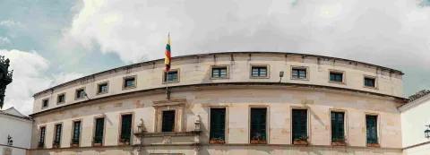 Vista panorámica de la fachada de un edificio histórico de estilo colonial con forma curva, construido en piedra clara. En el centro ondea la bandera de Colombia sobre la entrada principal, y a lo largo de la fachada se alinean ventanas con marcos oscuros y jardineras con flores. El cielo está parcialmente nublado, aportando un ambiente sobrio y patrimonial.