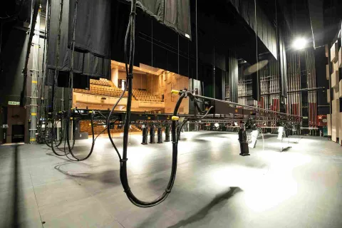  Vista desde el escenario de un teatro hacia el auditorio, donde se observan las graderías con asientos y la infraestructura técnica del escenario. En primer plano aparecen cables, estructuras de iluminación y sistemas de montaje escénico colgando del techo.