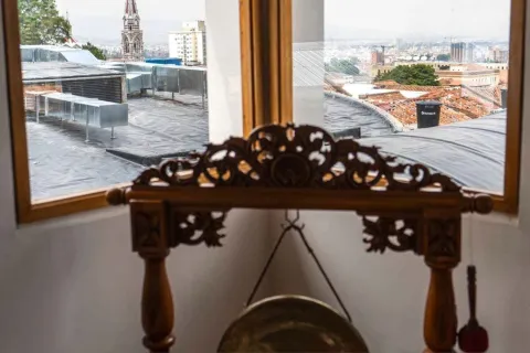  Interior de una habitación con una mesa decorativa de madera tallada y un pequeño gong colgante. Al fondo, a través de dos ventanas, se observa una vista del centro de la ciudad con techos de teja, edificios y una torre de iglesia sobresaliendo en el paisaje urbano.