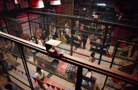 Vista elevada de un amplio espacio de eventos industrial con estructuras de malla metálica y una gran multitud de asistentes.
