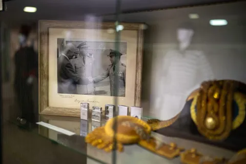  Vitrina de museo con objetos históricos de carácter militar o naval. En el centro se observa una fotografía en blanco y negro enmarcada, donde varios oficiales estrechan la mano, acompañada de pequeños objetos metálicos exhibidos en fila. En primer plano, desenfocado, se aprecia un elemento dorado decorativo, mientras la iluminación tenue resalta el valor histórico de la escena.