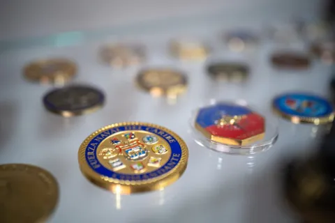 Primer plano de una vitrina con medallas y monedas conmemorativas de estilo militar o naval. En el centro destaca una pieza dorada con detalles en azul y escudos, mientras otras insignias se distribuyen alrededor, algunas dentro de estuches transparentes.