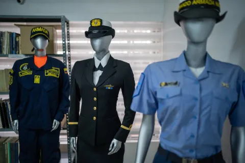 Tres maniquíes exhiben uniformes de la Armada en un espacio interior tipo museo. En el centro destaca un uniforme formal oscuro con botones dorados y gorra, mientras a los lados se observan otros trajes: uno azul con insignias y otro de tono claro con gorra. Al fondo hay estanterías con libros y una ventana con persianas que ilumina suavemente la escena.
