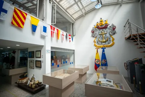  Interior de un museo con temática naval, iluminado por luz natural desde el techo. Se observan vitrinas de madera con documentos y objetos históricos, banderines marítimos colgados en la parte superior y, en una pared, un gran escudo con ancla y la frase “Plus Ultra”. A un lado están las banderas de Colombia y otra institucional, junto a una escalera metálica que conduce a un nivel superior.