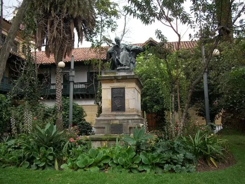 Estatua sobre pedestal en un jardín rodeado de árboles y plantas frente a una casa de estilo tradicional.