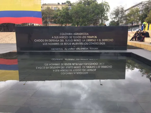  Monumento con placa conmemorativa a héroes caídos, reflejada en una superficie de agua, con bandera de Colombia al fondo.