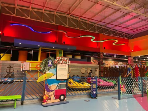 Atracción de carros chocones en un parque de entretenimiento interior, con luces de neón y vehículos de colores dentro de una pista cerrada.
