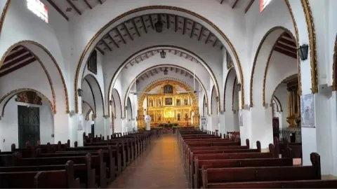 La imagen muestra el interior de una iglesia de estilo colonial, caracterizada por su amplitud y simetría. Un largo pasillo central de piso en tonos cálidos conduce la mirada hacia el altar mayor, mientras a ambos lados se alinean filas de bancas de madera oscura, ordenadas y silenciosas, que refuerzan la sensación de recogimiento y solemnidad.