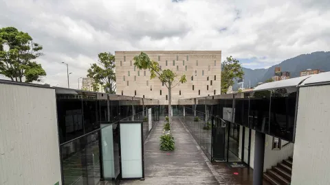 La imagen muestra un conjunto arquitectónico moderno con un corredor central de madera rodeado por estructuras de vidrio oscuro. Al fondo se levanta un edificio de piedra con ventanas verticales irregulares, mientras en el centro un árbol y algunas plantas aportan vida al espacio.