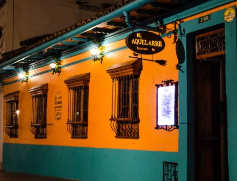 Fachada nocturna de un restaurante-bar de estilo colonial, pintado en tonos amarillo y turquesa. En la parte superior cuelga un letrero iluminado que dice “El Aquelarre”, acompañado de una escoba decorativa. La pared tiene varias ventanas con rejas de hierro y faroles encendidos que aportan una luz cálida.