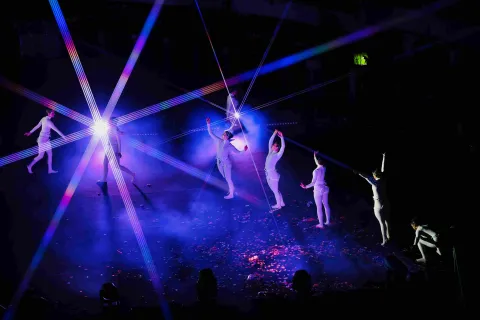  Escena teatral con varios bailarines vestidos de blanco sobre un escenario oscuro, iluminados por luces de colores y rayos láser que cruzan el espacio. Algunos performers están de pie y otros en movimiento, rodeados de humo y confeti en el suelo.