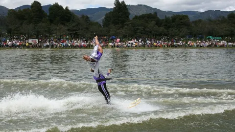 Persona practicando esquí acuático en un lago, realizando una acrobacia frente a espectadores en la orilla.