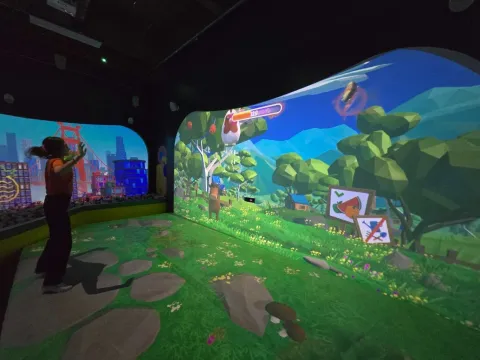 Niños jugando frente a una pantalla interactiva con paisaje desértico animado, rodeados de cubos de colores en un espacio digital inmersivo.