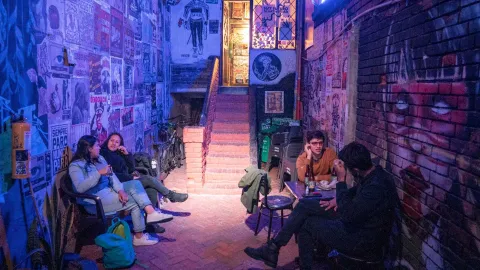 Interior de un bar moderno con iluminación ambiental en tonos verdes y azules. En la pared izquierda se lee “Hanna Hops Brewery” en letras grandes, mientras que al fondo un letrero neón dice “Be the brewer!”. En el centro, dos personas están sentadas en una mesa redonda conversando, rodeadas de bancas de madera y plantas decorativas.