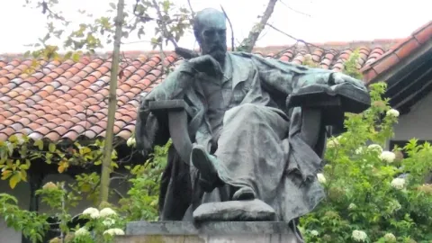 Escultura de un hombre sentado sobre pedestal, rodeada de vegetación y con techo de tejas al fondo.