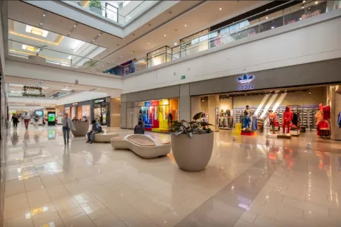  La imagen muestra el interior de un centro comercial moderno y elegante, con amplios pasillos iluminados por luz natural que entra desde el techo. A un lado se observa una tienda de ropa con vitrinas coloridas y maniquíes, mientras que en el centro hay mobiliario de descanso y una gran maceta con plantas.