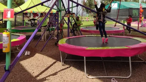 Persona salta en un trampolín con arnés de seguridad en un parque al aire libre, mientras otras personas preparan el equipo alrededor.