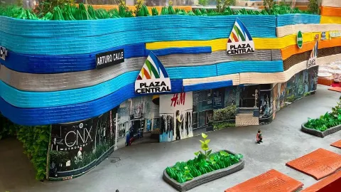 La imagen presenta una maqueta detallada y colorida de un centro comercial identificado como ´Plaza Central'. La fachada destaca por su diseño ondulado y vibrante, compuesta por franjas en distintos tonos de azul, amarillo, naranja y blanco, que le dan un carácter moderno, dinámico y atractivo.