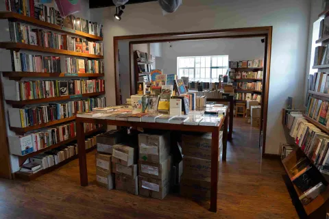  Interior de una librería con estanterías llenas de libros, una mesa central de exhibición y cajas de ejemplares apiladas debajo.