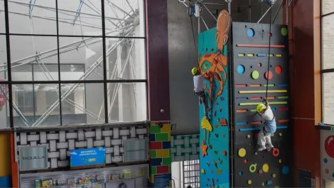 Dos niños con casco escalan un muro de escalada interior con paneles coloridos y figuras decorativas.