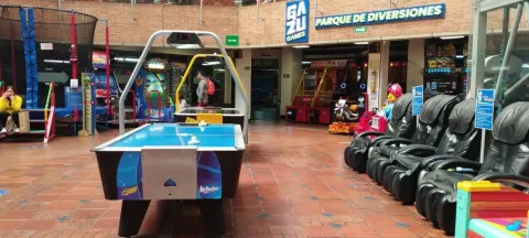 Zona de juegos en un centro comercial con mesa de air hockey, máquinas arcade y sillas de masaje, bajo un letrero de parque de diversiones.