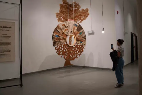 Persona observa y fotografía una obra artística colgante con forma de árbol en una sala de museo.