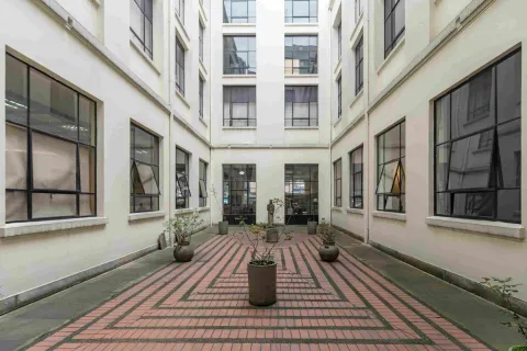 La imagen muestra un patio interior de estilo sobrio y ordenado, rodeado por paredes claras y grandes ventanas. En el centro, varias macetas con plantas se distribuyen sobre un piso de ladrillo dispuesto en patrones geométricos, creando un ambiente tranquilo y equilibrado.