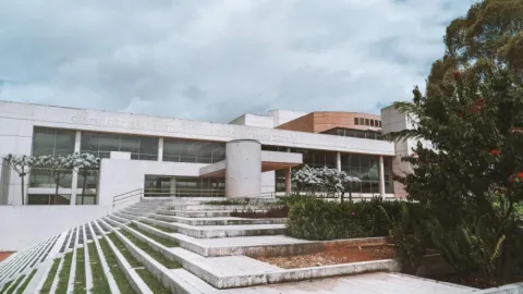 Modern cultural building with wide staircases, large windows, and gardens under a cloudy sky.