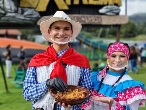 Dos personas con trajes tradicionales sonríen y muestran una canasta con comida típica en un parque al aire libre.
