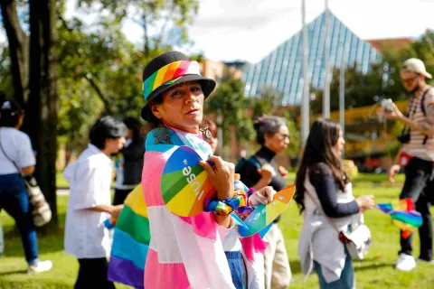 Persona en un parque durante una marcha del orgullo LGBTI+, con sombrero negro adornado con una cinta arcoíris y una bandera multicolor sobre los hombros; sostiene abanicos con colores de la diversidad mientras otras personas caminan alrededor en un ambiente soleado.