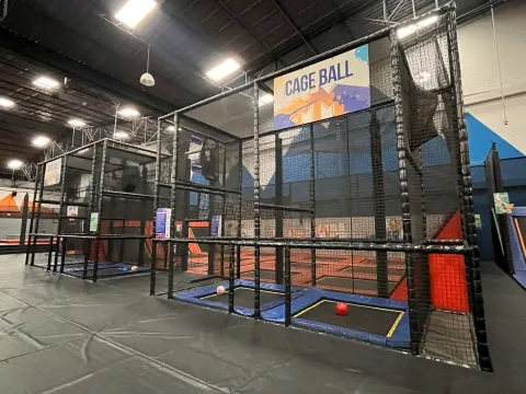Área de juego “cage ball” en un parque interior, con canchas enrejadas, trampolines y pelotas dentro de una estructura cerrada.