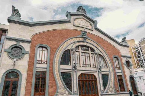 La imagen muestra la fachada de un edificio histórico con detalles ornamentales y estilo clásico, donde destacan los arcos decorativos y el trabajo en piedra y ladrillo. En el centro, una gran entrada enmarcada por formas curvas y elementos artísticos resalta su carácter cultural.