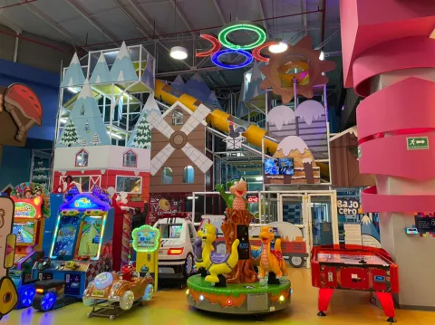 Área de juegos infantiles en un parque interior con máquinas arcade, figuras coloridas y una gran estructura de juegos con toboganes y decoraciones temáticas.