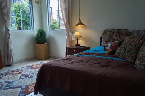 La imagen muestra una habitación acogedora y bien iluminada, con una cama vestida en tonos cálidos y cojines coloridos. La luz natural entra por dos ventanas con cortinas claras, complementada por una lámpara de mesa que aporta calidez.