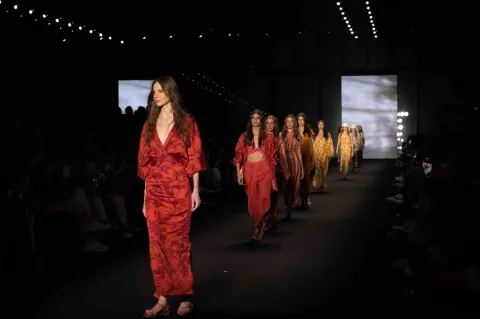 Fila de modelos desfila en pasarela con vestidos en tonos rojos y estampados, ante público en un evento de moda.